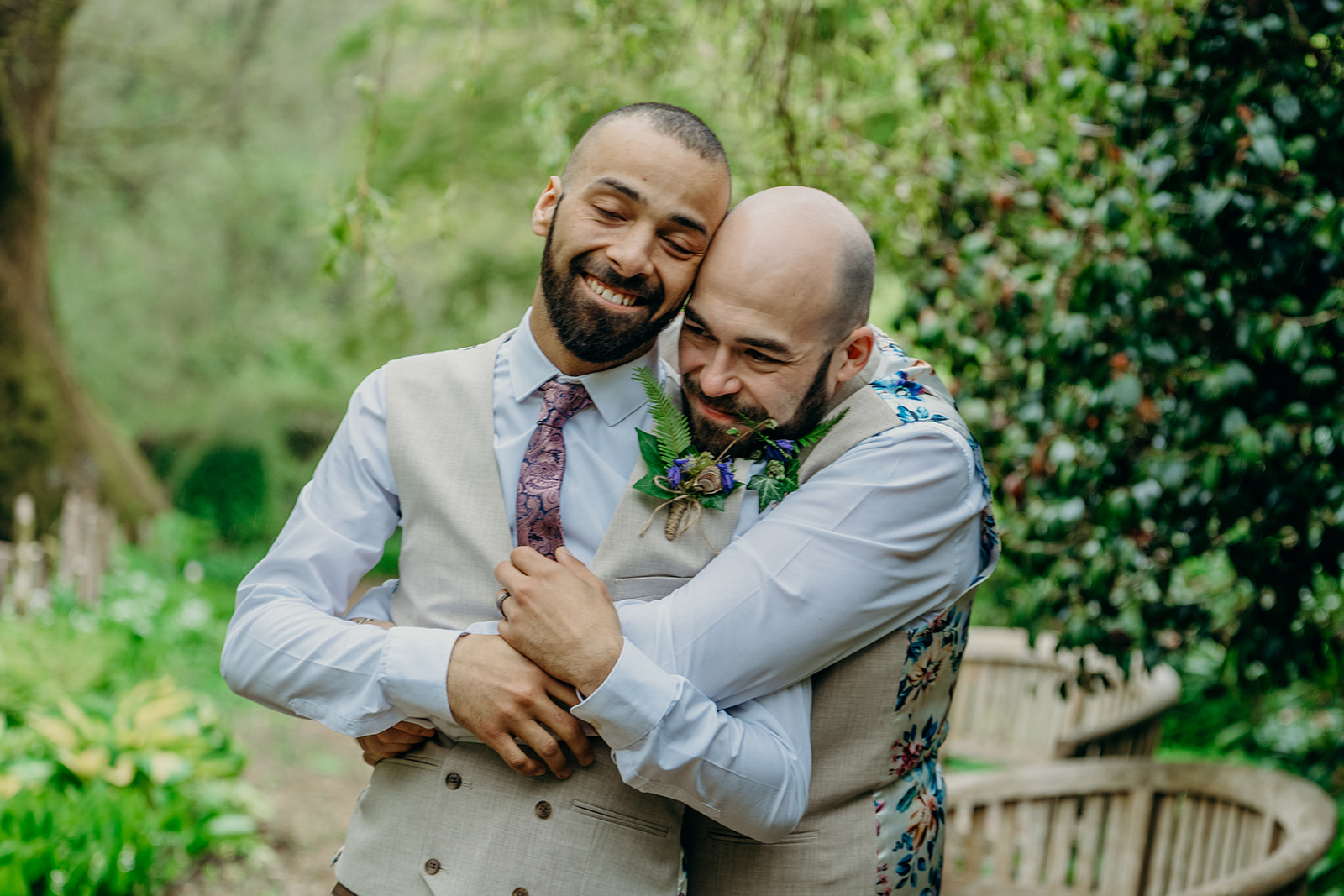 two shaven headed bearded grooms dressed in matching cream linen waistcoats with flowery backs hugging outdoors in a garden