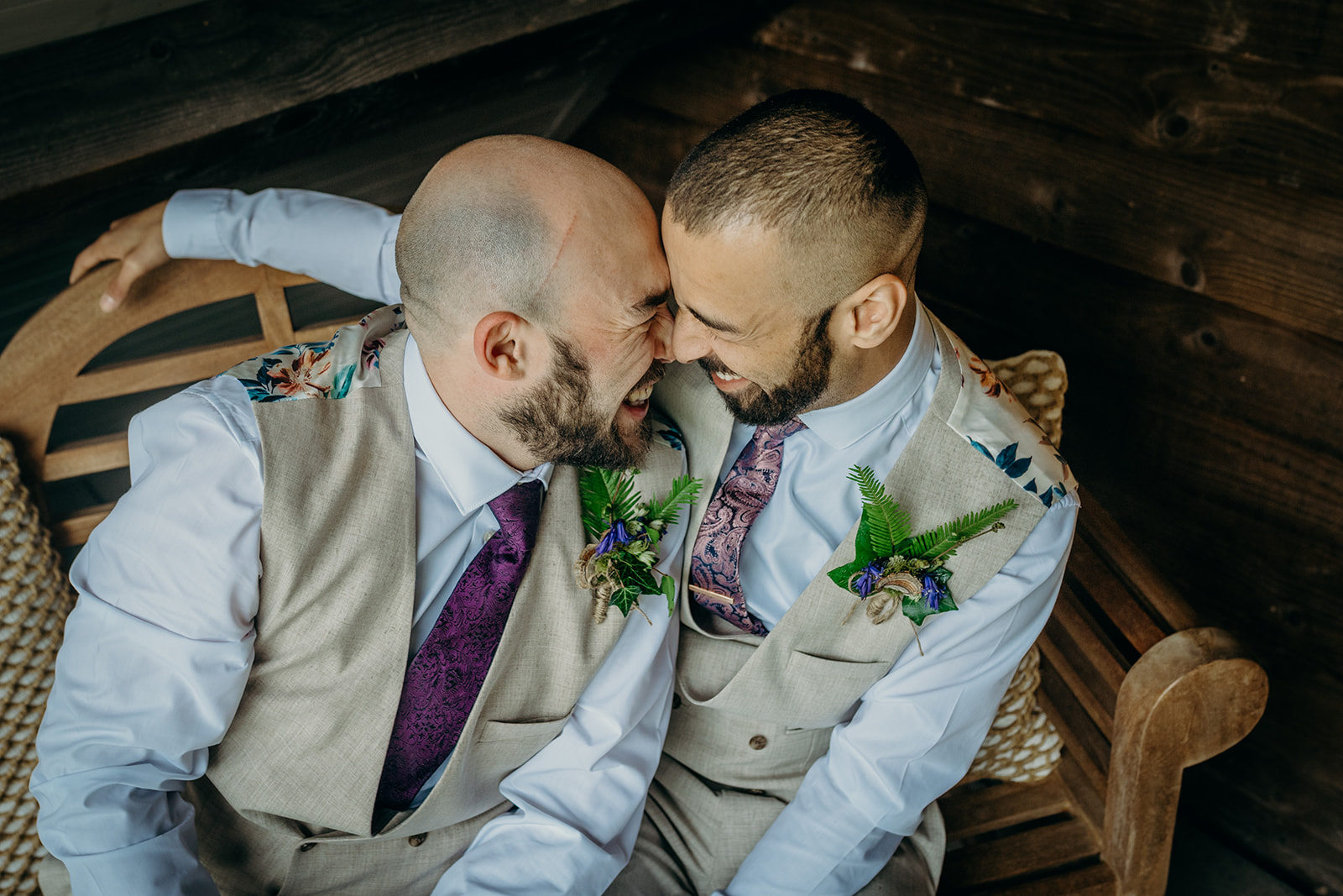 two shaven headed bearded groom in cream linen waistcoats purple neckties and eleborate fern boutonniers sat on an oak bench smiling with faces touching