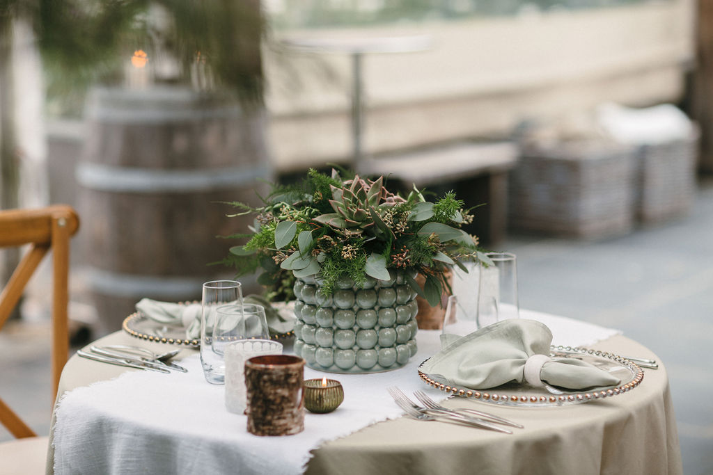 foreground shot of wedding table set for two with sage green bobble vase and succulent bouquet and rustic decor in background