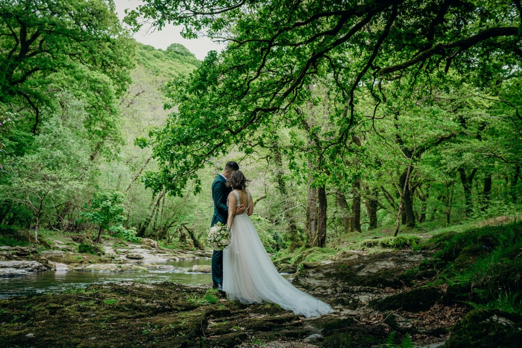 back view of wedding couple stood on river bank surrounded by green trees