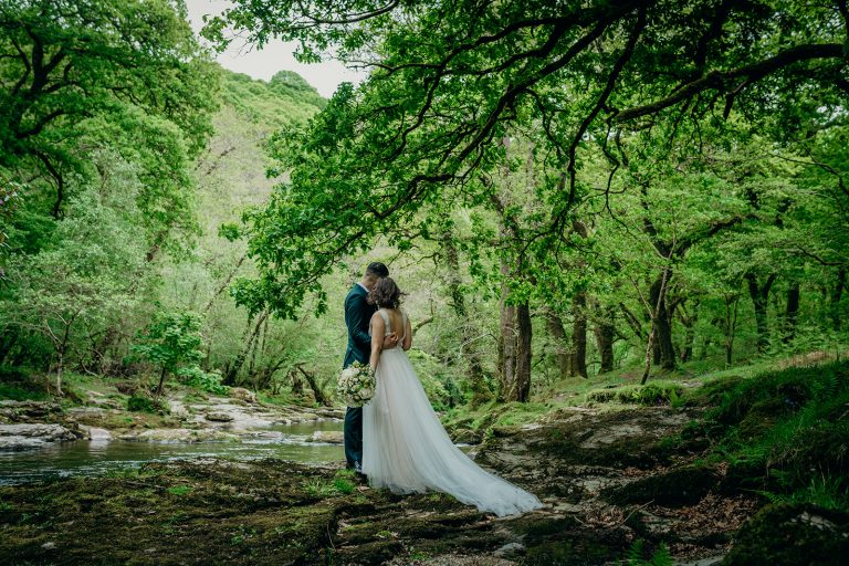 rear shot of bride and groom on the banks of a river surrounded by green trees