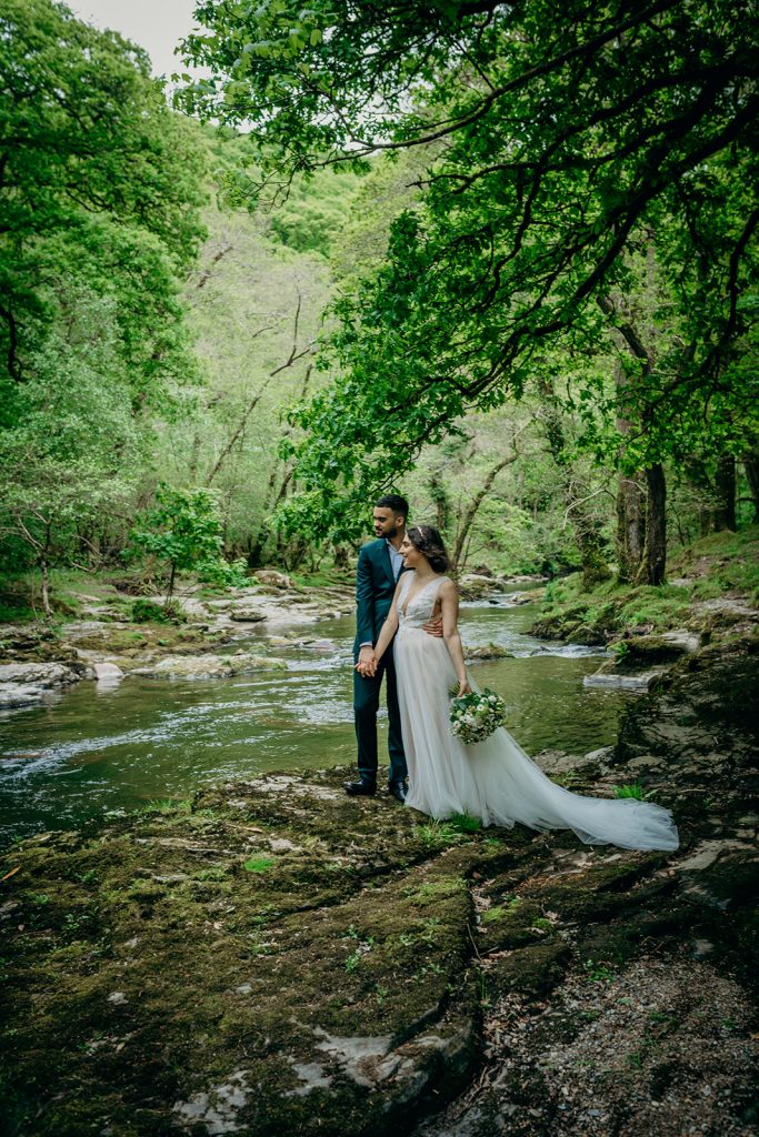 bride in tulle wedding gown holding bouquet stood with groom on banks of a Dartmoor river