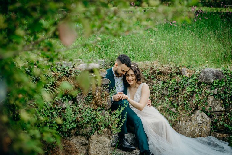 bride wearing tulle wedding dress sat with groom on stone wall smiling