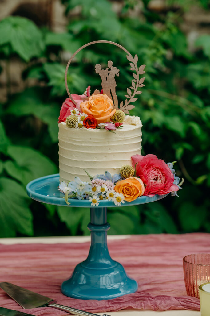 small single tier wedding cake in rough buttercream icing on a blue glass cake stand decorated with brightly coloured flowers
