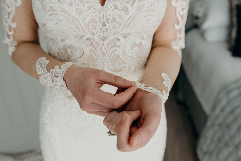 bride doing up the bottom button of her sleeve on her wrist. detail of the lace on her dress.