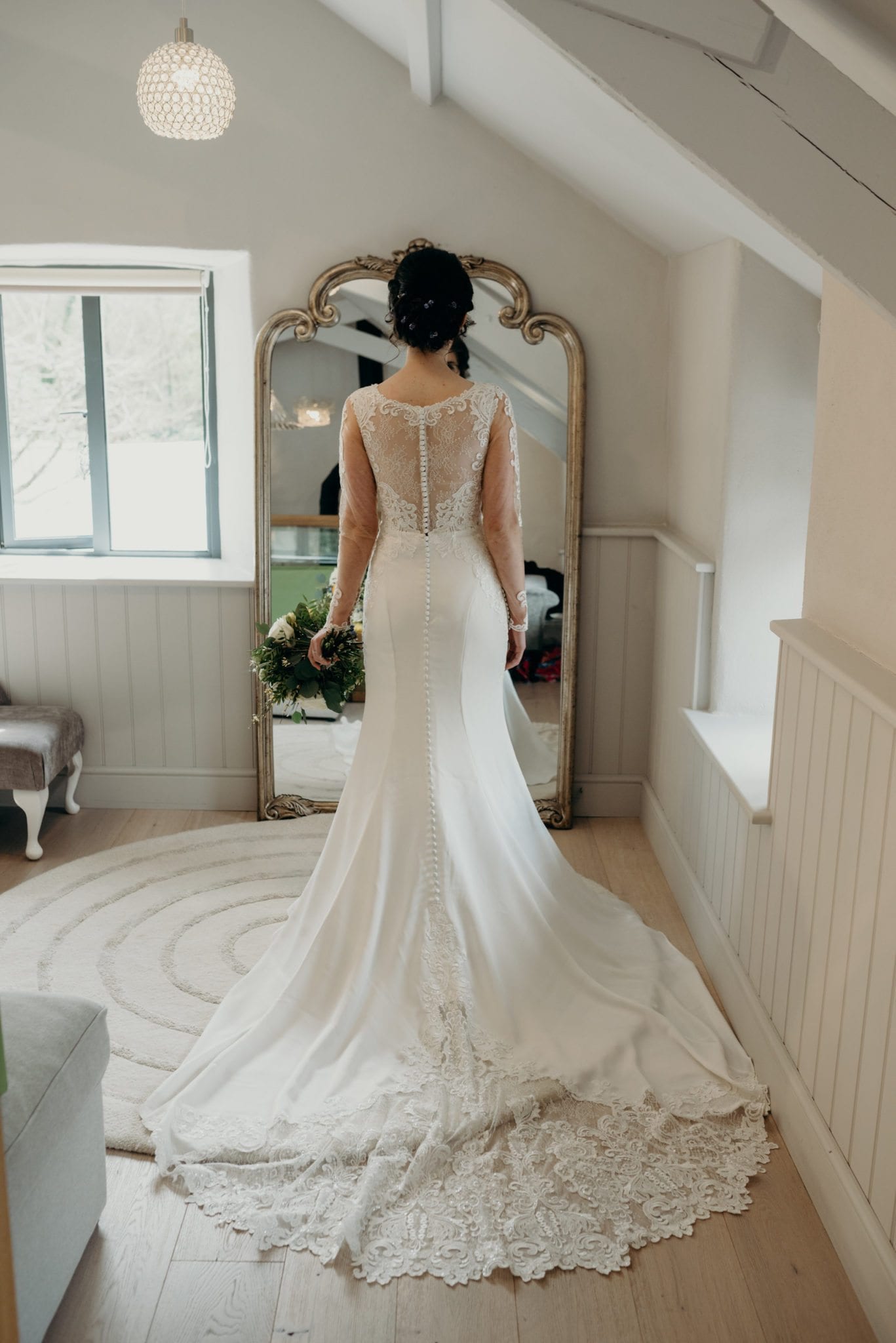 bride standing in mirror with bouquet to her left. in focus is her mirage image; lace dress with long sleeves, v neck, and a plan skirt. Slightly out of focus is the back view of the dress with illusion lace and buttons.