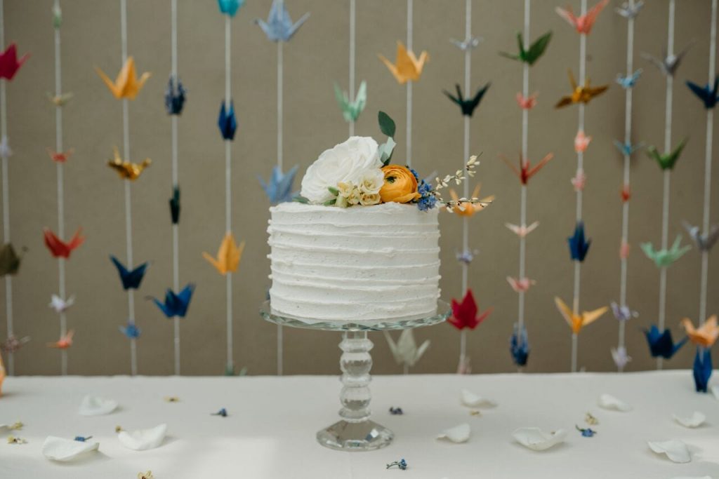 miniature elopement wedding cake with ivory buttercream icing decorated with ivory, bright orange and navy blue flowers on a backdrop of colourful hanging paper cranes