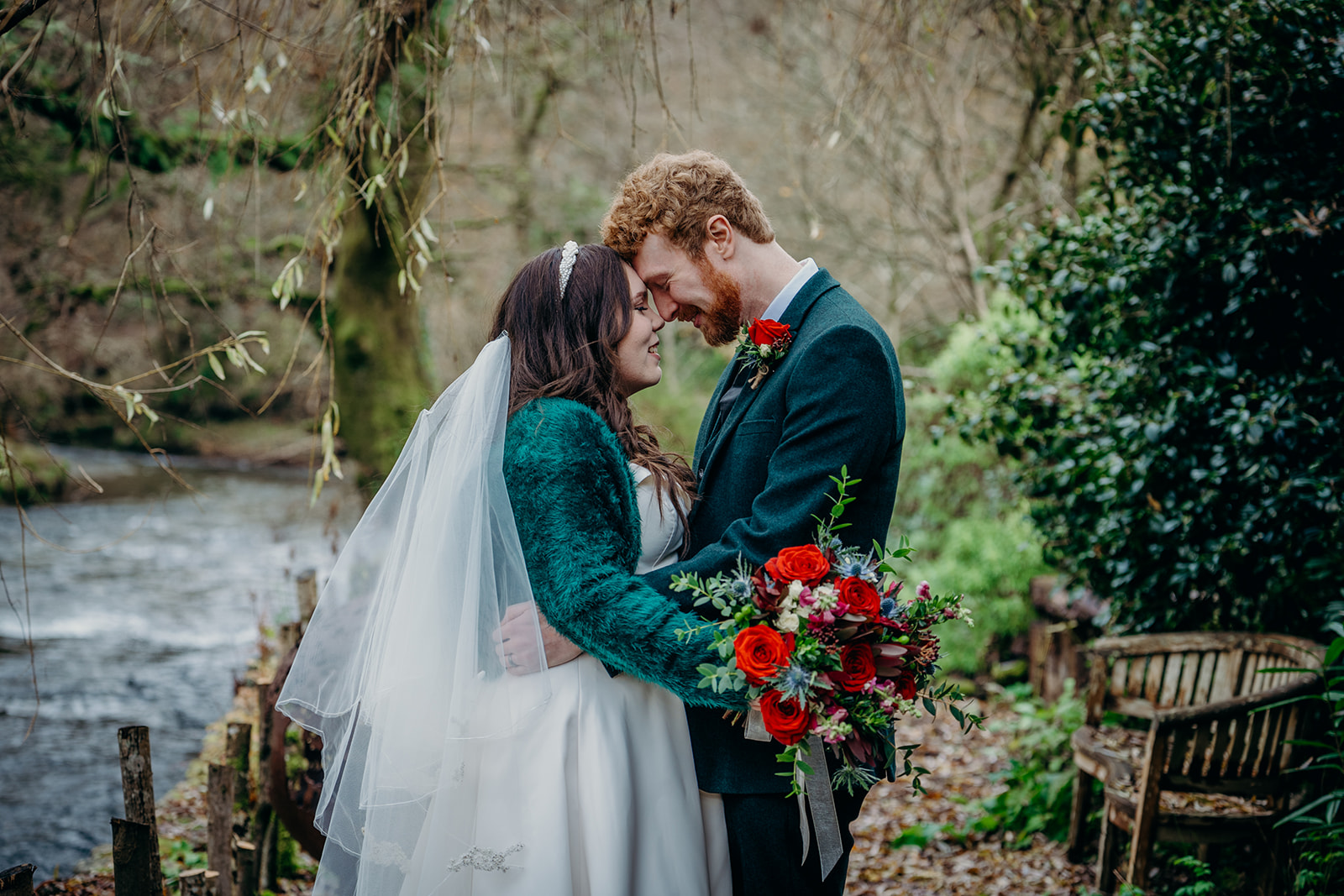 bride and groom in dark green outfits on the banks of a river with red rose bouquet and buttonhole